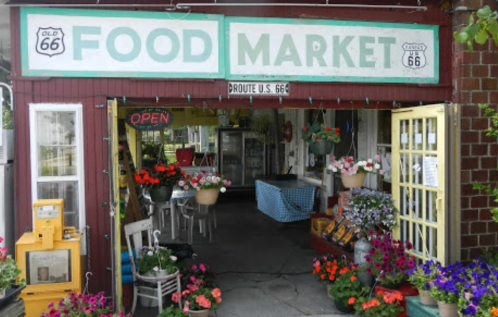 A charming food market entrance adorned with flowers, featuring a sign that reads "Open" and "Route U.S. 66."