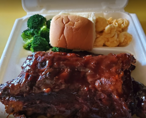 A takeout container with BBQ ribs, broccoli, a roll, and macaroni and cheese.