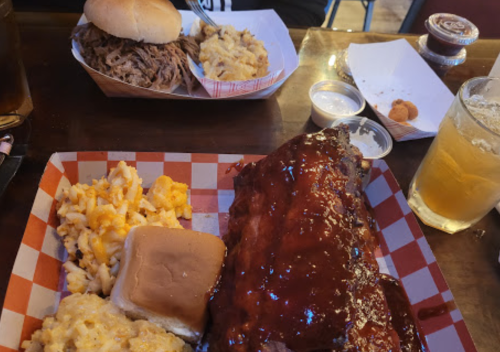 A plate of BBQ ribs with mac and cheese, a bun, and a drink, alongside a pulled pork sandwich and more mac and cheese.