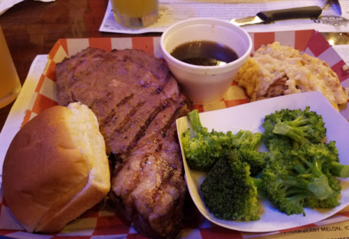 A plate featuring grilled steak, a bun, broccoli, macaroni and cheese, and a cup of sauce on a red-checkered tray.