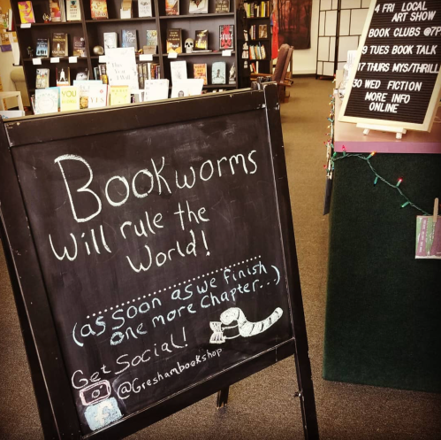 A chalkboard sign in a bookstore reads, "Bookworms will rule the world! Get social!" with a playful design.