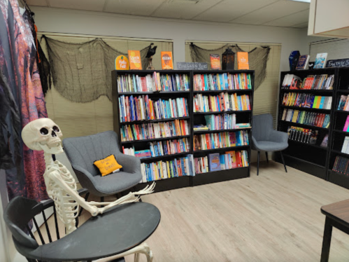 A cozy reading room with bookshelves, a skeleton sitting on a chair, and Halloween decorations.