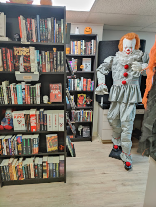 A spooky room with a clown figure and a bookshelf filled with horror books, including works by Stephen King.