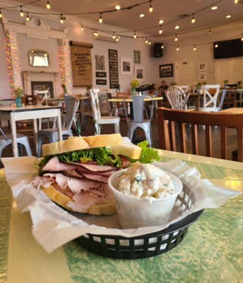 A sandwich with turkey and lettuce on a plate, alongside a small cup of potato salad, in a cozy café setting.