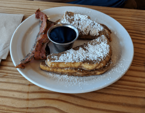 A plate with three slices of French toast dusted with powdered sugar, a side of bacon, and a small cup of syrup.