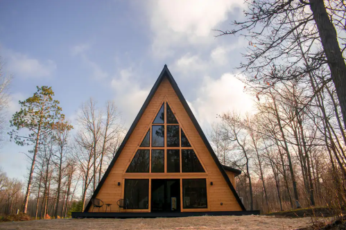 A modern A-frame cabin surrounded by trees under a cloudy sky. Large windows reflect the natural setting.