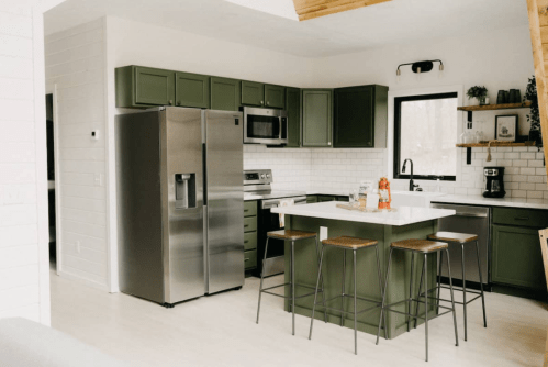 Modern kitchen with green cabinets, stainless steel appliances, and a white island with bar stools. Bright and airy design.