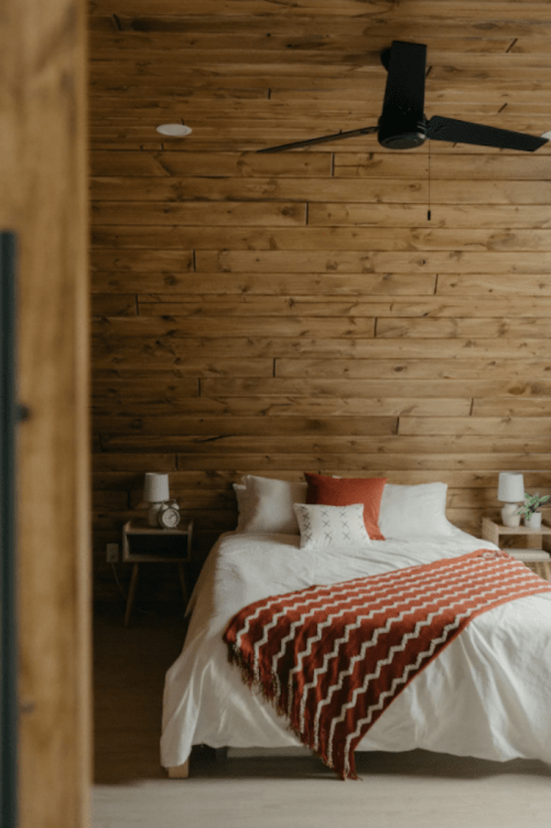 Cozy bedroom with a wooden wall, a bed adorned with a red and white blanket, and stylish bedside lamps.