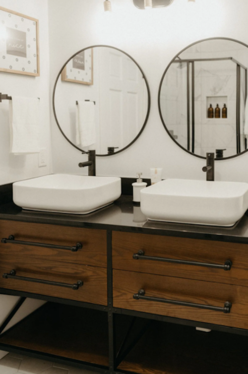 Modern bathroom with two round mirrors, white sinks, and a wooden vanity with black accents.