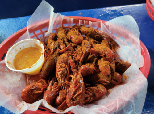 A red basket filled with seasoned crawfish and a small cup of dipping sauce on a blue table.