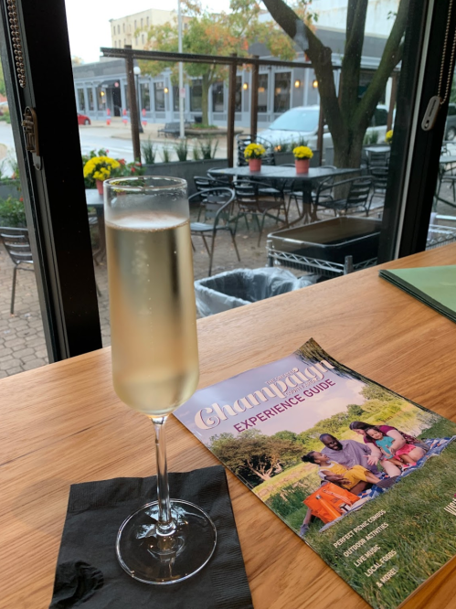 A glass of champagne on a wooden table next to a magazine titled "Champaign Experience Guide," with outdoor seating visible.