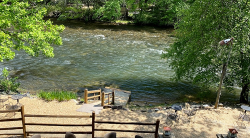 A serene river flows through a lush green landscape, with a wooden dock and seating area by the water's edge.