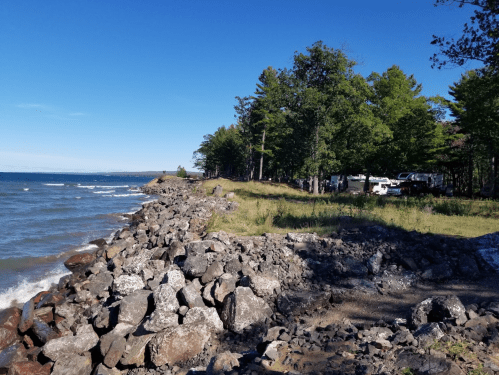 A rocky shoreline with trees and a clear blue sky, overlooking a calm lake.