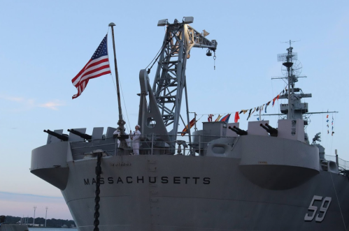 A naval ship named Massachusetts with an American flag and colorful flags on display against a clear sky.