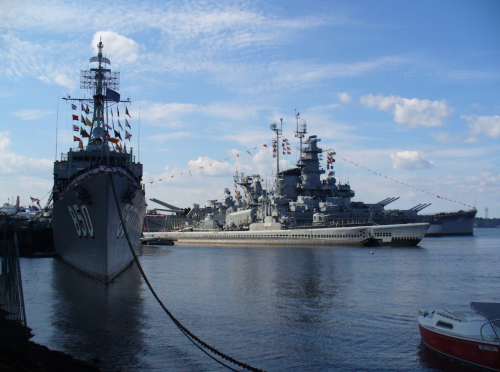 Two large naval ships docked by the waterfront under a blue sky with scattered clouds.