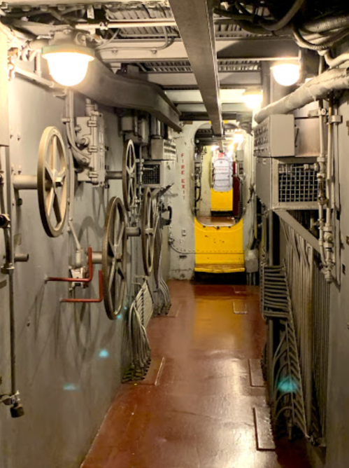 A narrow, industrial hallway with metal walls, pipes, and wheels, leading to a yellow door at the far end.
