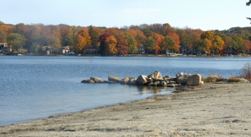A serene lake scene with colorful autumn trees, rocky shoreline, and calm water under a clear sky.