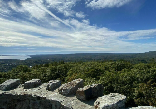 A scenic view from a rocky overlook, showcasing lush green trees and a vast sky with wispy clouds.
