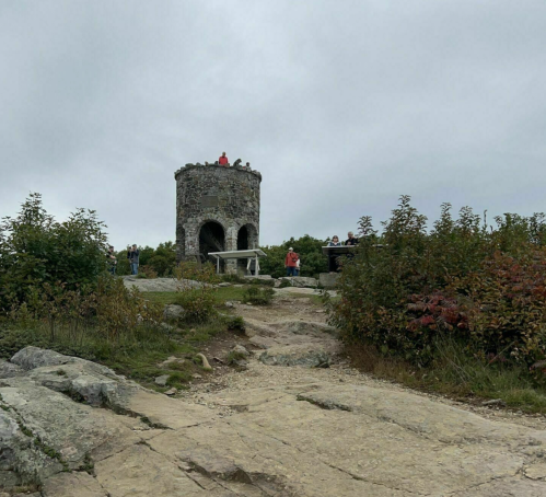 A stone tower on a rocky path, surrounded by greenery and people, under a cloudy sky.