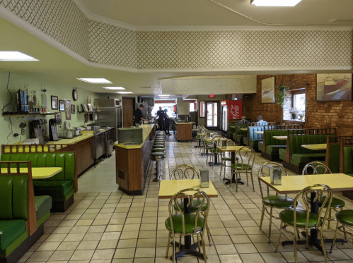 A retro diner interior with green booths, yellow tables, and a tiled floor, featuring a counter and vintage decor.