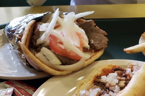 A close-up of a sandwich filled with sliced meat, onions, and tomatoes, wrapped in foil on a plate.