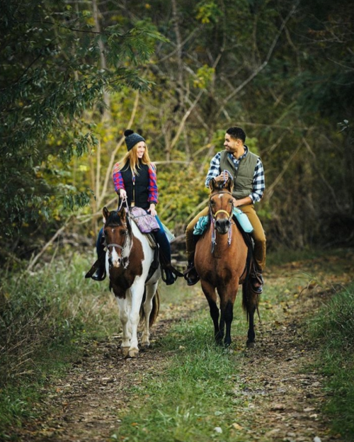 A man and woman ride horses along a wooded trail, smiling and enjoying their time outdoors.