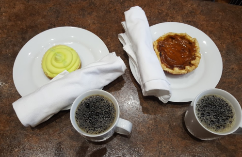 Two plates with a green swirl dessert and a caramel tart, accompanied by two cups of coffee on a brown table.