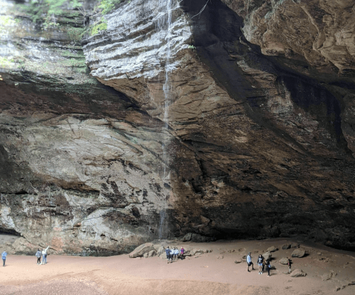 A group of people stands beneath a large rock overhang with a waterfall cascading down in a natural cave setting.