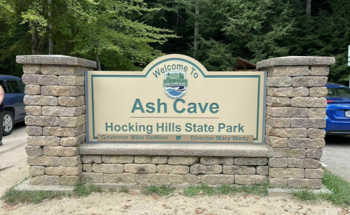 Welcome sign for Ash Cave at Hocking Hills State Park, featuring stone pillars and greenery in the background.
