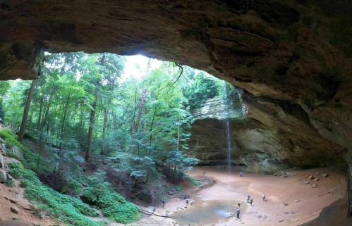 A panoramic view of a cave with a sandy floor, surrounded by lush green trees and a waterfall in the background.