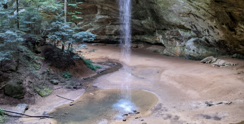 A serene waterfall cascades into a sandy area surrounded by lush greenery and rocky cliffs.