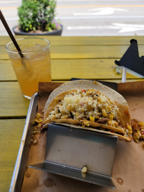 A taco filled with meat and toppings on a tray, with a drink in the background on a yellow table.