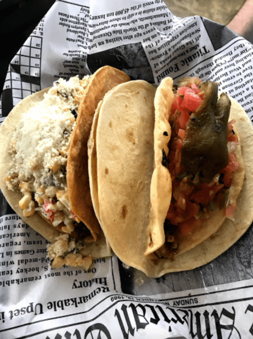 Three tacos on a plate, filled with various ingredients like meat, cheese, and fresh toppings, wrapped in newspaper.