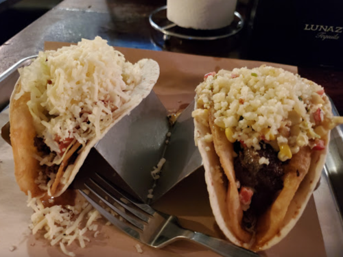 Two tacos on a tray, filled with meat, topped with cheese, lettuce, and diced tomatoes, with a fork beside them.