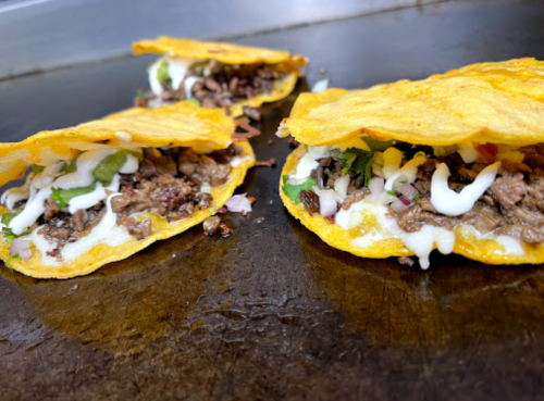 Three tacos filled with beef, cheese, onions, and cilantro, served on a griddle.