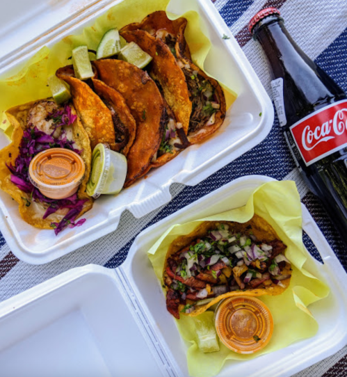 A takeout container with tacos, lime wedges, and a bottle of Coca-Cola on a striped tablecloth.
