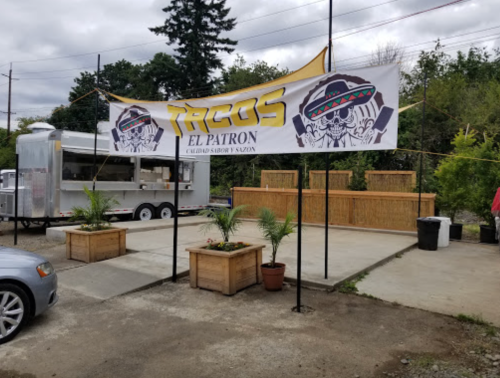 A food truck with a banner reading "Tacos El Patron" in a casual outdoor setting with planters and a cloudy sky.