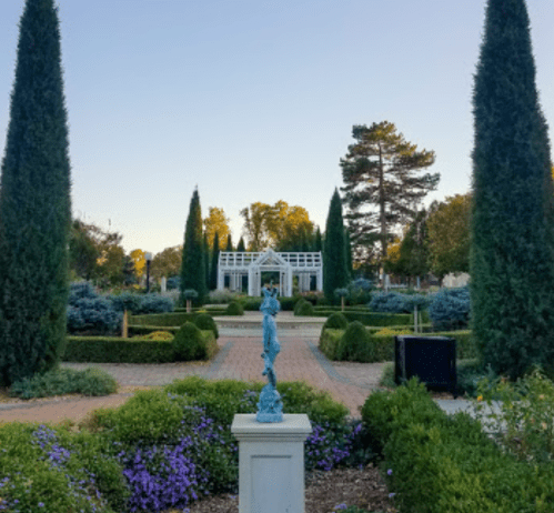 A serene garden with tall conifer trees, a statue in the foreground, and a white gazebo in the background.