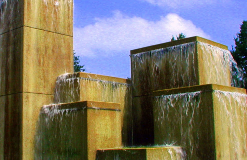 A modern stone fountain with cascading water, surrounded by greenery and a blue sky.