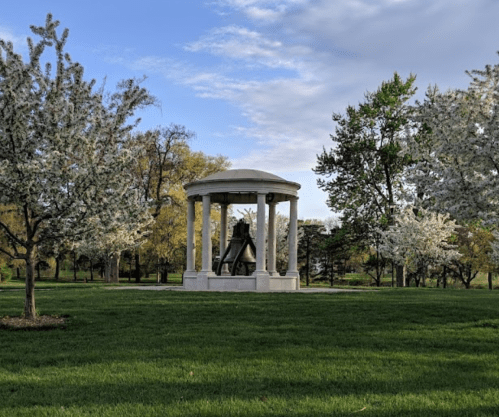 A white gazebo surrounded by blooming trees in a green park under a partly cloudy sky.