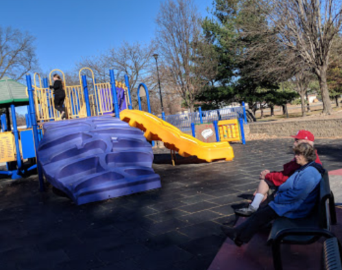 A playground with a slide and climbing structure, two people sitting on a bench nearby, and trees in the background.