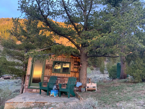 A cozy cabin nestled among trees, featuring two green chairs on a porch and a scenic mountain backdrop.