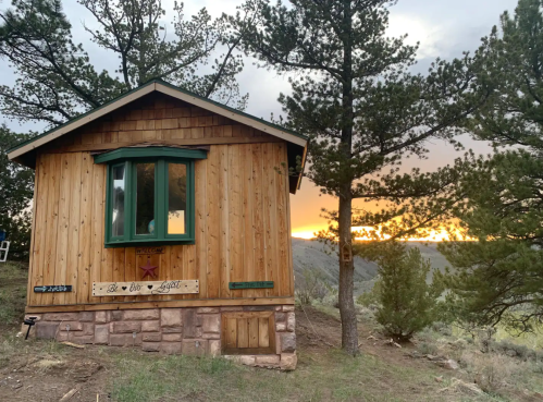 A wooden cabin surrounded by pine trees, with a sunset glowing in the background.