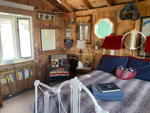 Cozy bedroom with wooden walls, a bed with blue linens, a reading chair, and various decorative items.