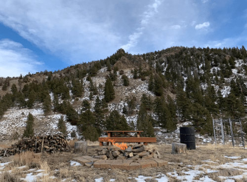 A fire pit surrounded by logs, set against a snowy mountain landscape with pine trees under a blue sky.