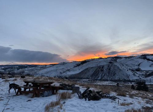 A snowy landscape at sunset, with mountains in the background and a picnic table in the foreground.