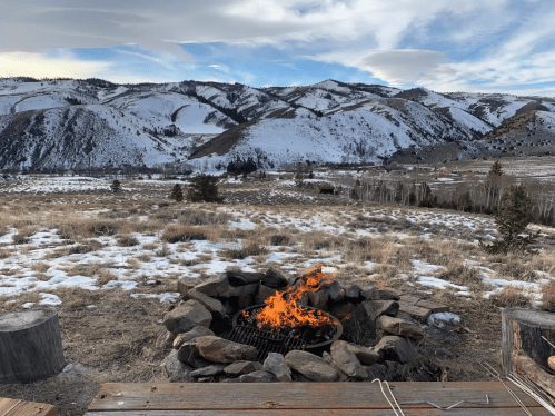 A stone fire pit with flames, set against a snowy mountain landscape under a cloudy sky.
