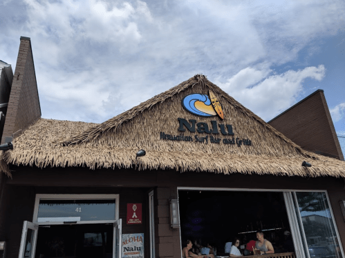 A thatched-roof building with a sign reading "Nalu Hawaiian Surf Bar and Grill" under a cloudy sky.