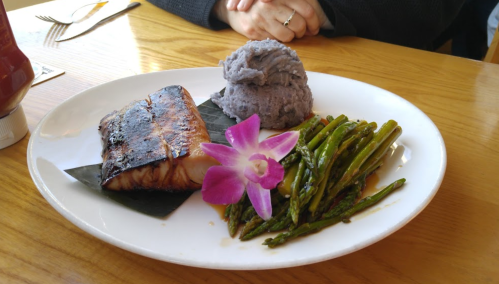 A plate with grilled fish, purple mashed potatoes, asparagus, and a flower garnish on a wooden table.