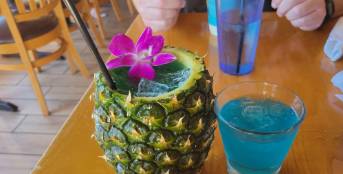 A pineapple drink garnished with a flower, next to a blue cocktail on a wooden table.